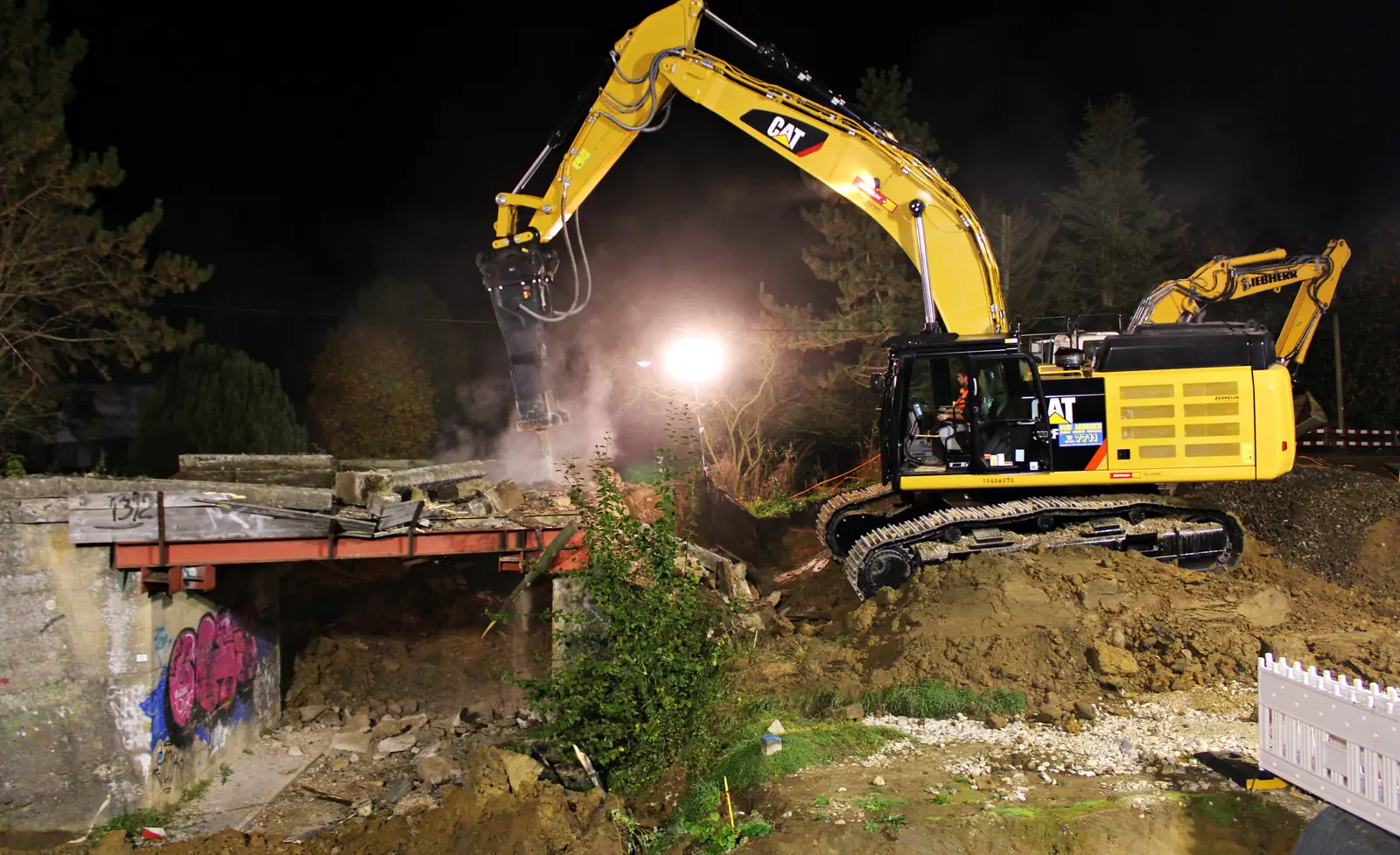 Bub-Bagger mit Abbruchzange an einer Bahnbrücke in der Nacht, Flutlicht und Staub
